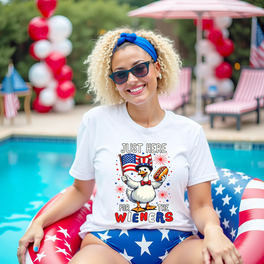 Female model wearing white ‘Just Here for the Wieners’ t-shirt outdoors, cute goose with flag and hot dog graphic, fun July 4th BBQ vibe