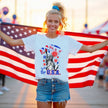 Female model wearing white ‘Party in the USA’ Statue of Liberty t-shirt outdoors, Liberty holding red cup design with fireworks and patriotic summer vibe.