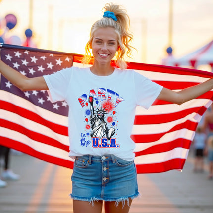 Female model wearing white ‘Party in the USA’ Statue of Liberty t-shirt outdoors, Liberty holding red cup design with fireworks and patriotic summer vibe.
