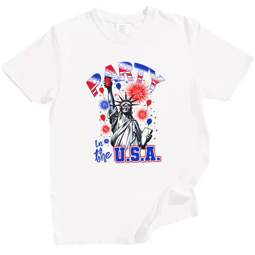 Female model wearing white ‘Party in the USA’ Statue of Liberty t-shirt outdoors, Liberty holding red cup design with fireworks and patriotic summer vibe.