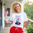 Female model wearing white ‘Party Like a Patriot’ t-shirt outdoors, George Washington bust with party hat and fireworks, fun July 4th party vibe.
