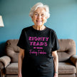 Woman wearing black ‘80 Years of Being Fucking Fabulous’ birthday t-shirt with bold pink mixed-font lettering, confident and playful 80th birthday celebration vibe.
