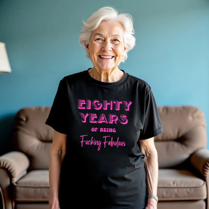 Woman wearing black ‘80 Years of Being Fucking Fabulous’ birthday t-shirt with bold pink mixed-font lettering, confident and playful 80th birthday celebration vibe.