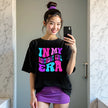 Woman wearing ‘In My Birthday Girl Era’ black birthday t-shirt outdoors, bold retro pink, purple, and blue lettering, playful and confident birthday celebration vibe