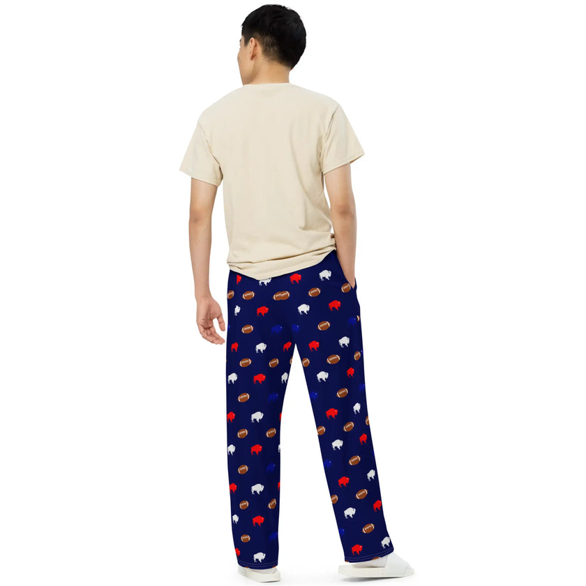 Unisex navy pajama pants with repeating Buffalo football pattern featuring red white and blue buffalo silhouettes and brown footballs in relaxed wide leg design as seen from behind on male model.