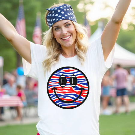 Blonde mom wearing white Buffalo football t-shirt with red white and blue striped smile face graphic and football eyes at outdoor festival in park setting, modern Buffalo game day apparel.