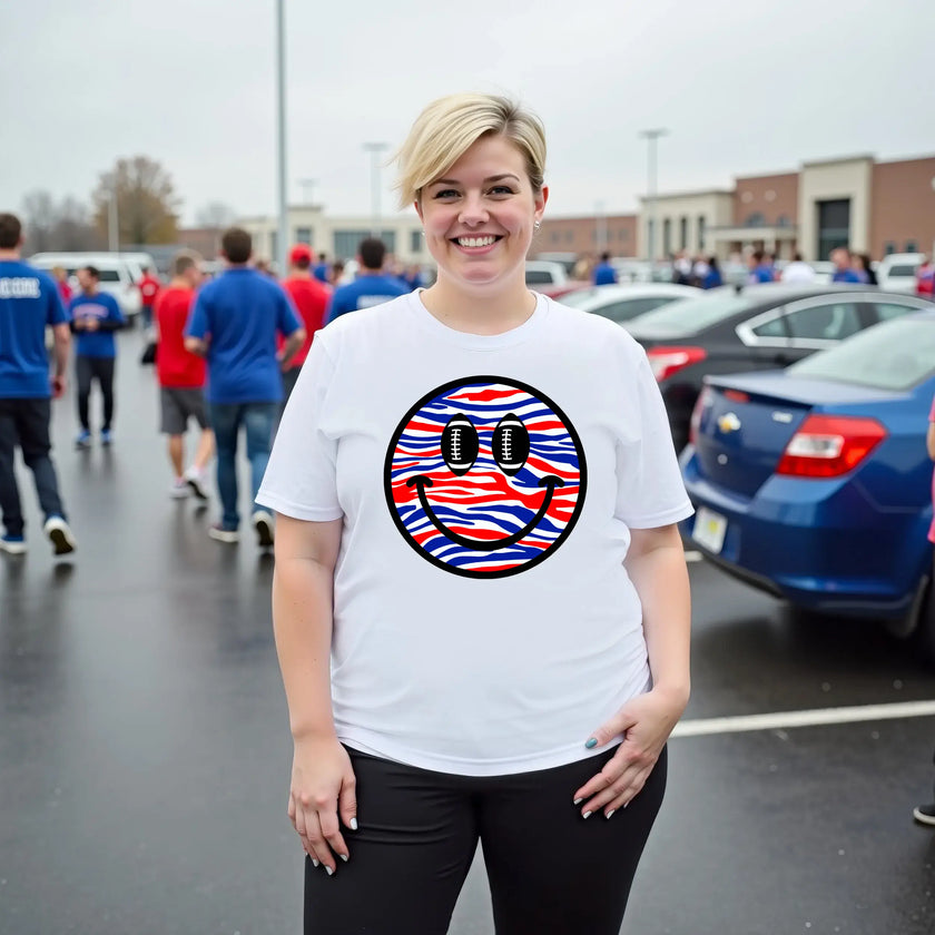 Female model wearing white Buffalo football smiling face t-shirt outdoors, red white blue striped design with football eyes, relaxed game day vibe.