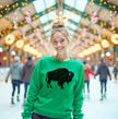 A smiling blonde woman stands on an indoor ice rink wearing a bright green crewneck sweatshirt with a large black buffalo graphic on the front. The background shows a festive, blurred ice rink with lights and people skating.