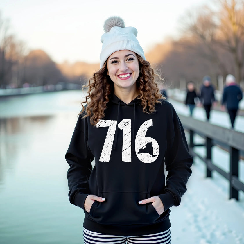 Female model wearing black Buffalo NY 716 hoodie in the snow, distressed white numbers with New York state cutout, cozy winter local pride look.