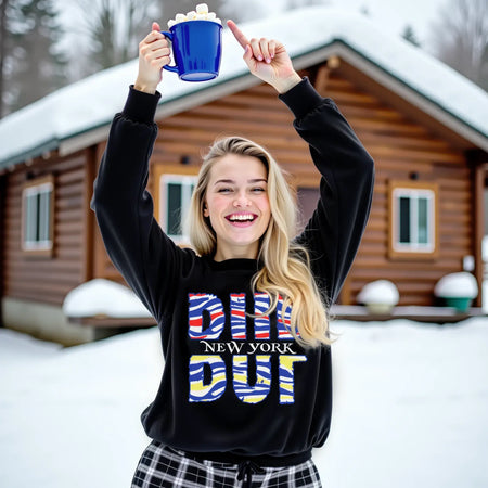 young woman wearing a black buffalo ny football hockey hybrid sweatshirt outside at the cabin in the snow. she is holding a cup of hot cocoa up and pointing to it.