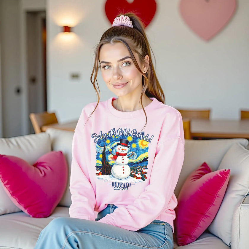 Female model wearing pink Buffalo winter snowman sweatshirt sitting on couch in cozy living room setting