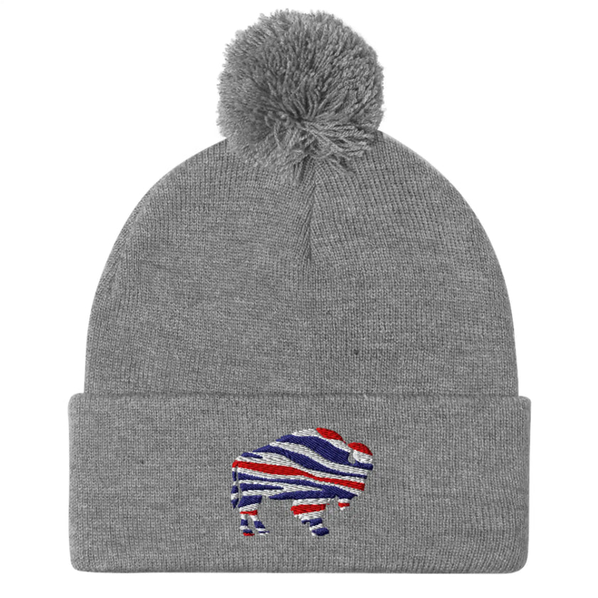 Grey puffball beanie featuring embroidered Buffalo silhouette with red white blue striped football team colors, cozy Buffalo winter hat.