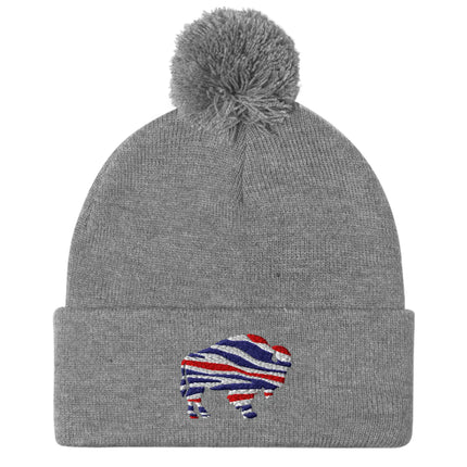Grey puffball beanie featuring embroidered Buffalo silhouette with red white blue striped football team colors, cozy Buffalo winter hat.