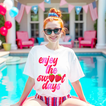 Female model wearing white Enjoy the Sweet Days strawberry t-shirt in pool, retro distressed lettering with fruit detail, cute summer vibe.