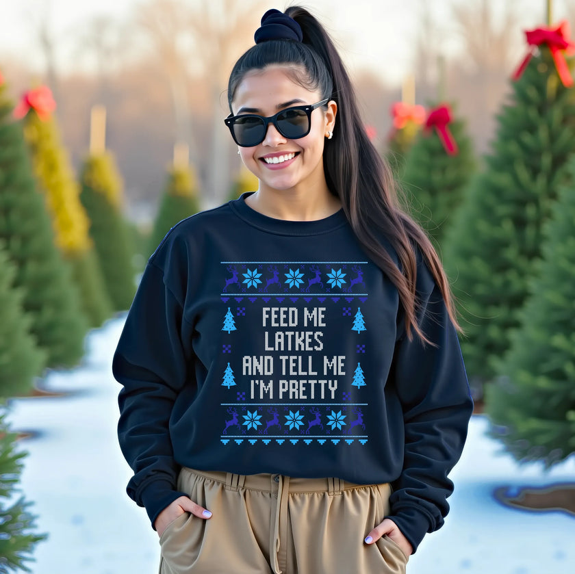 Female model wearing navy Hanukkah ugly sweater style sweatshirt featuring ‘Feed Me Latkes and Tell Me I’m Pretty’ knitted-look lettering with festive reindeer and snowflake design, cozy cute holiday winter apparel.
