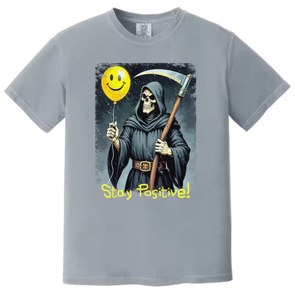 Gray “Stay Positive” grim reaper t-shirt flat lay showcasing dark humor artwork with scythe and cheerful balloon contrast