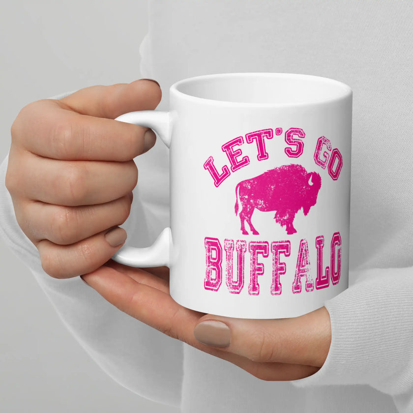 Hand holding white ceramic coffee mug featuring hot pink distressed ‘Lets Go Buffalo’ Buffalo football design with buffalo silhouette, game day drinkware.
