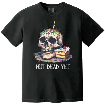 Black “Not Dead Yet” t-shirt flat lay showcasing skull with lit birthday candle, rainbow sprinkles, and cake slice, funny dark humor birthday design