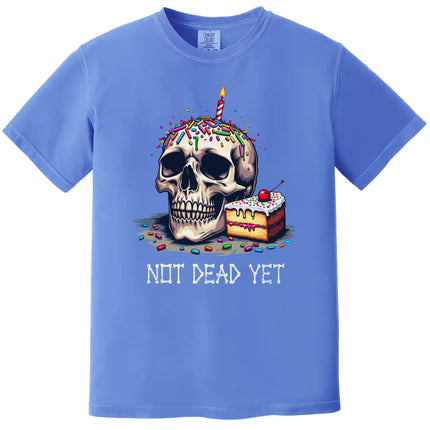 Blue “Not Dead Yet” t-shirt flat lay featuring birthday skull with candle, sprinkles, and cake slice, playful sarcastic graphic tee