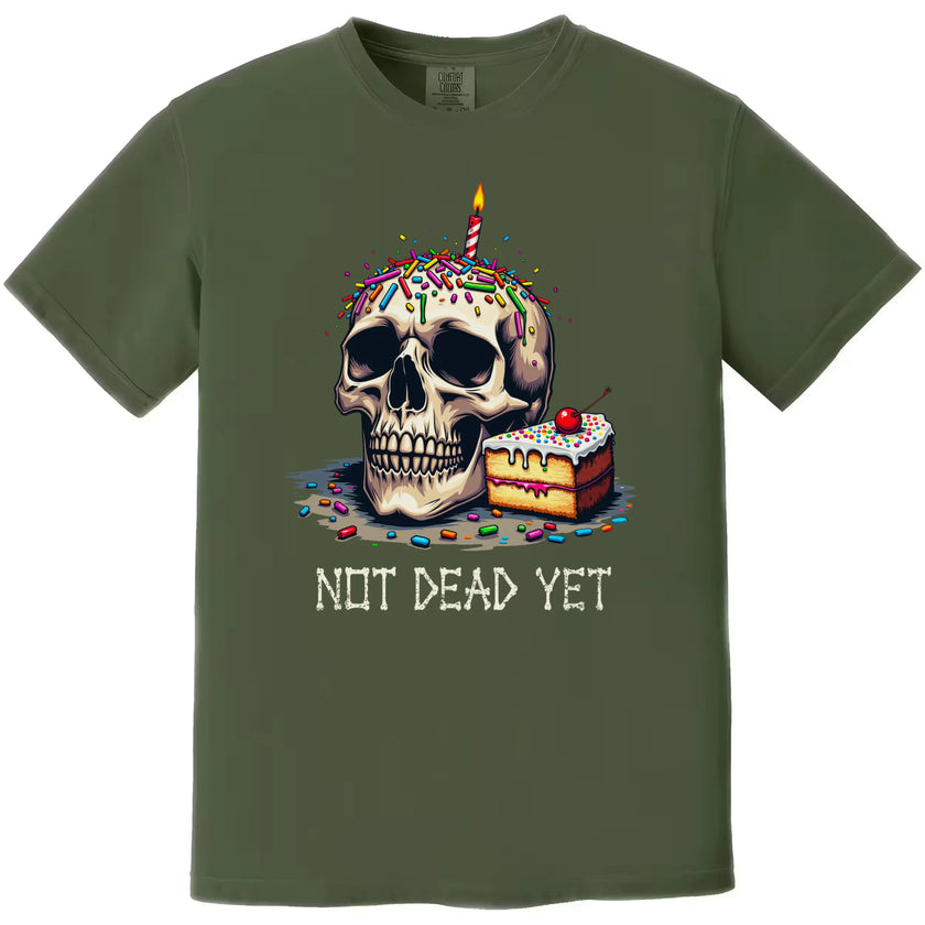Green “Not Dead Yet” t-shirt flat lay with skull, candle, rainbow sprinkles, and cake, sarcastic birthday design for milestone celebrations