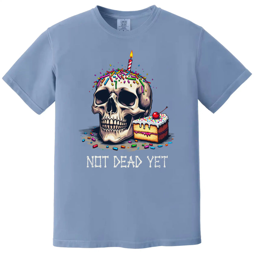 Light blue “Not Dead Yet” t-shirt flat lay showcasing skull with lit candle, rainbow sprinkles, and birthday cake slice, funny and dark humor tee
