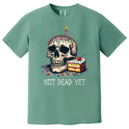 Green “Not Dead Yet” t-shirt flat lay with skull and lit birthday candle, rainbow sprinkles, and cake slice, dark humor birthday tee