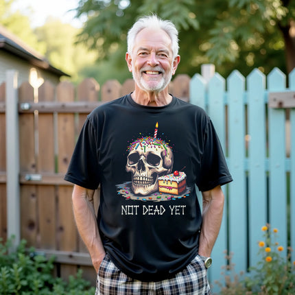 Model wearing black “Not Dead Yet” t-shirt in casual lifestyle setting, showing skull with lit birthday candle, rainbow sprinkles, and cake slice, sarcastic birthday vibe