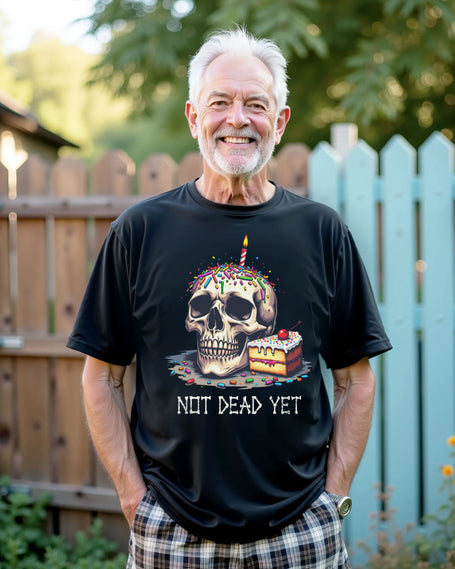 Older man standing in backyard wearing black ‘Not Dead Yet’ birthday t-shirt featuring skull with sprinkles, birthday candle, and cake slice graphic, cover image for birthday apparel collection.