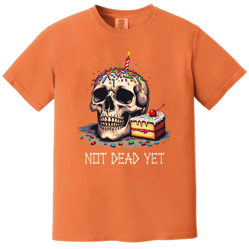 Orange “Not Dead Yet” t-shirt flat lay with skull, birthday candle, sprinkles, and cake slice, playful and sarcastic birthday graphic