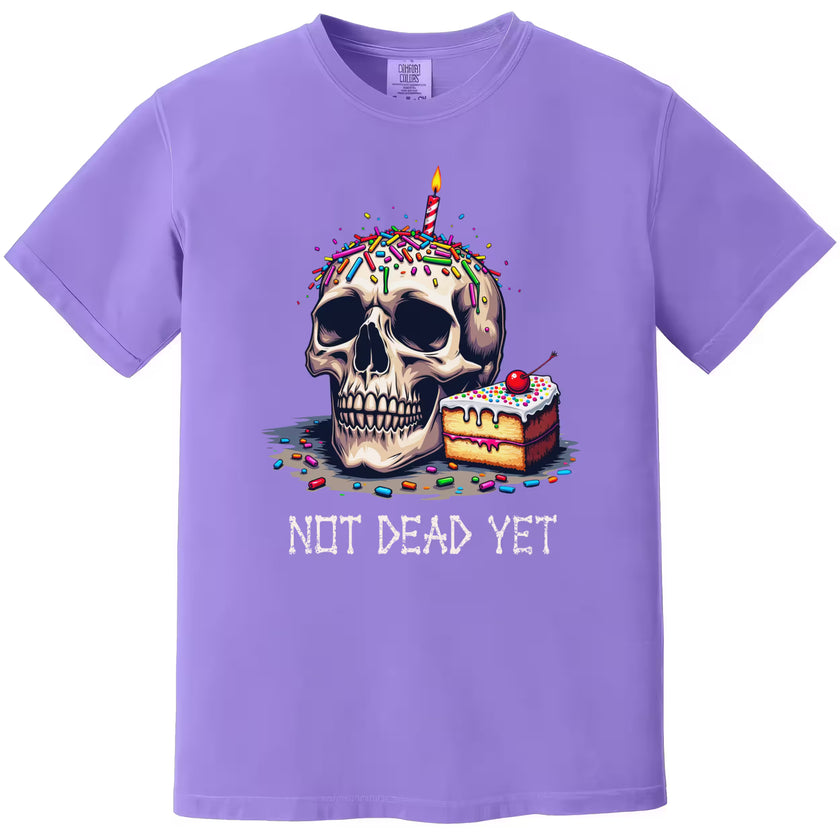Purple “Not Dead Yet” t-shirt flat lay featuring skull with lit candle, rainbow sprinkles, and cake slice, cheeky birthday humor design
