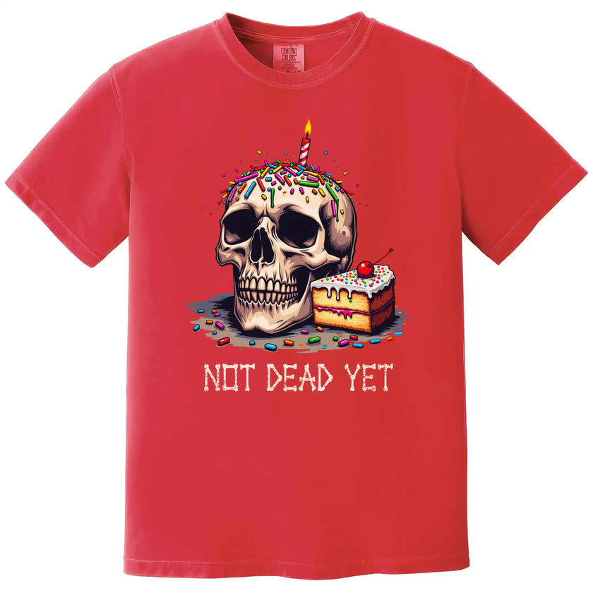 Red “Not Dead Yet” t-shirt flat lay featuring skull with lit birthday candle, rainbow sprinkles, and slice of cake, dark humor birthday design