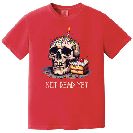 Red “Not Dead Yet” t-shirt flat lay featuring skull with lit birthday candle, rainbow sprinkles, and slice of cake, dark humor birthday design