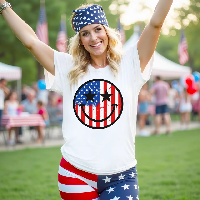 Female model wearing white smiling face patriotic t-shirt at park festival, paint splatter American flag design with star eyes, fun July 4th vibe.