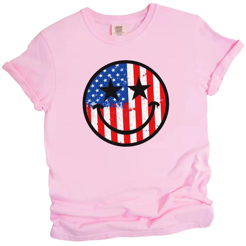 Cute patriotic smiling face shirt in pink, American flag paint splatter design with star eyes, playful July 4th tee.