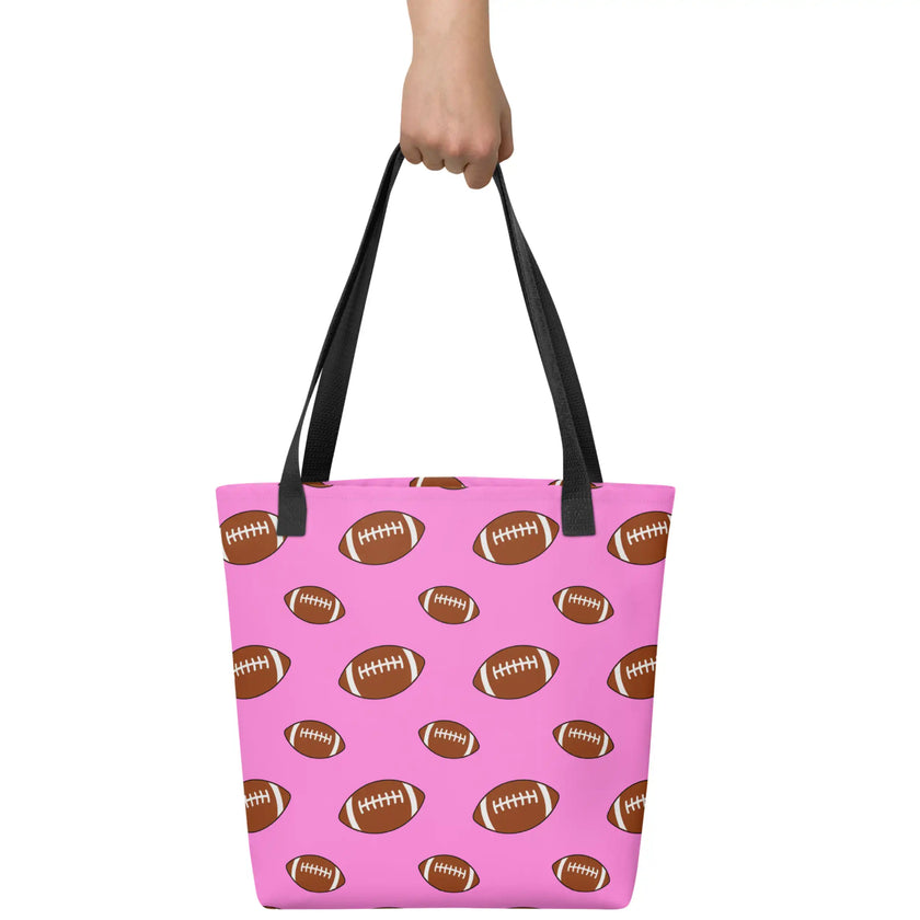 Hand holding a pink football print tote bag featuring repeating brown footballs in multiple sizes, versatile carryall for shopping, groceries, books, and travel.