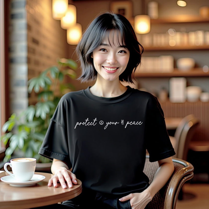 Female model wearing black “Protect Your Peace” t-shirt in a casual lifestyle setting, sitting comfortably and showing relaxed, intentional energy