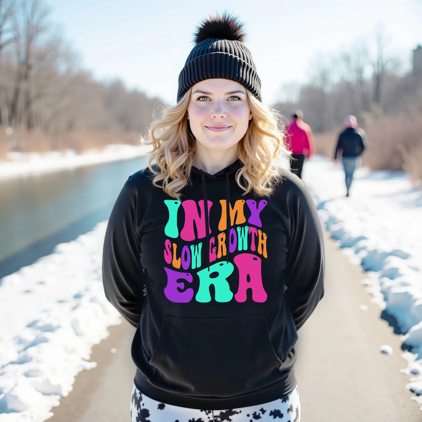 Female model wearing black “In My Slow Growth Era” hoodie in a casual lifestyle setting, cozy and confident look celebrating personal growth