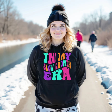 Female model wearing black “In My Slow Growth Era” hoodie in a casual lifestyle setting, cozy and confident look celebrating personal growth