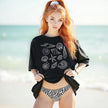 Female model wearing black seashell summer t-shirt on the beach, featuring detailed ocean shell illustrations in a minimalist coastal design.