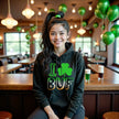 Female model wearing black Buffalo shamrock St. Patrick’s Day hoodie sitting inside an Irish pub, cozy and festive