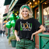 Female model wearing black Lucky Vibes St. Patrick’s Day t-shirt standing outside an Irish pub celebrating the holiday