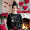 Female model wearing black gothic skull heart checkbox sweatshirt standing by a Valentines-decorated fireplace, showing playful dark-goth style