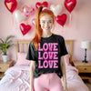 Female model wearing black ‘All We Need Is Love’ Valentine’s tee with pink lettering, styled standing cute in her bedroom