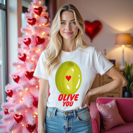 Female model wearing white Olive you funny pun t-shirt, standing in front of a pink Valentine’s tree in a living room, smiling.