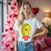 Female model wearing white Olive you funny pun t-shirt, standing in front of a pink Valentine’s tree in a living room, smiling.