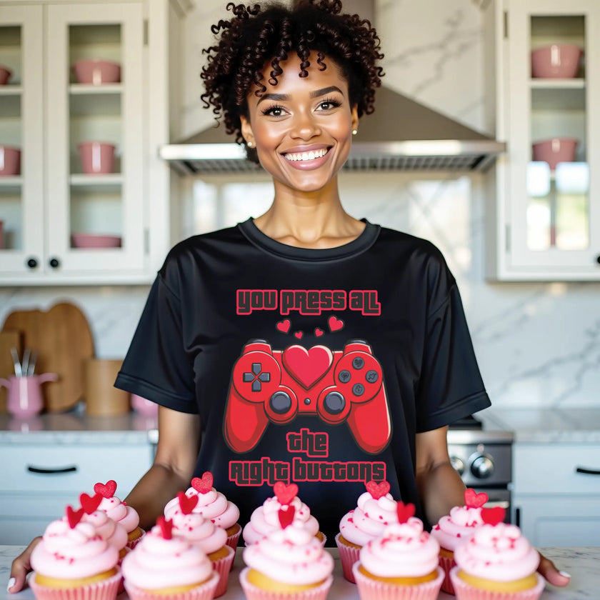 Female model wearing black Valentine’s gamer tee with red game controller and heart, standing in kitchen with cupcakes