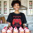 Female model wearing black Valentine’s gamer tee with red game controller and heart, standing in kitchen with cupcakes
