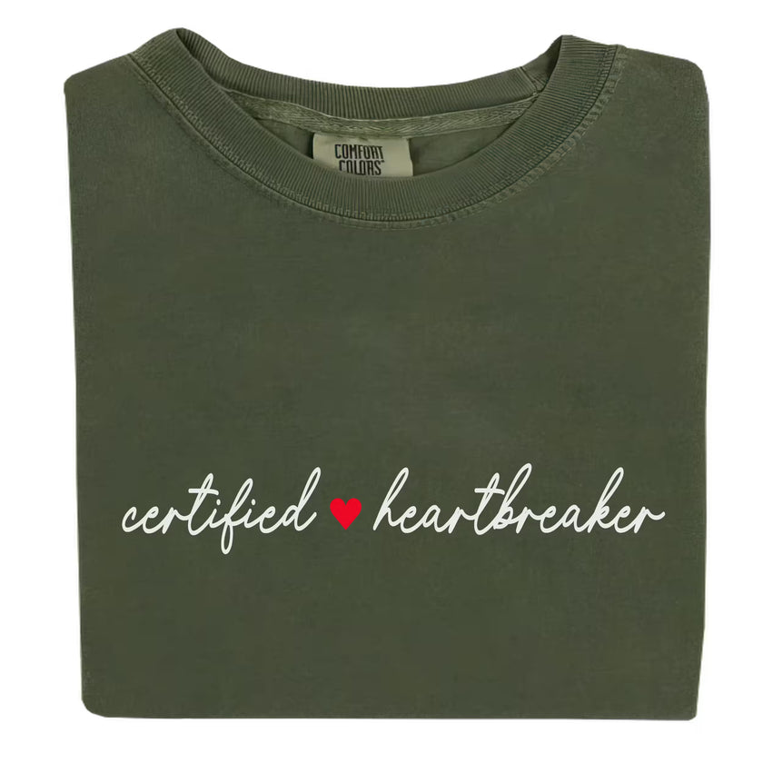 Folded flat lay of green Certified Heartbreaker tee with red heart graphic on white background.