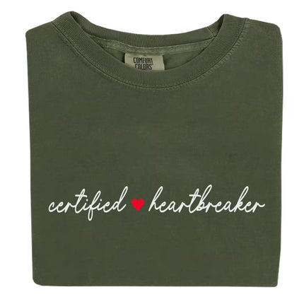 Folded flat lay of green Certified Heartbreaker tee with red heart graphic on white background.