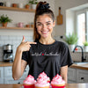 Female model wearing black Certified ♥ Heartbreaker tee in the kitchen, styled with cupcakes for a playful Valentine’s vibe.
